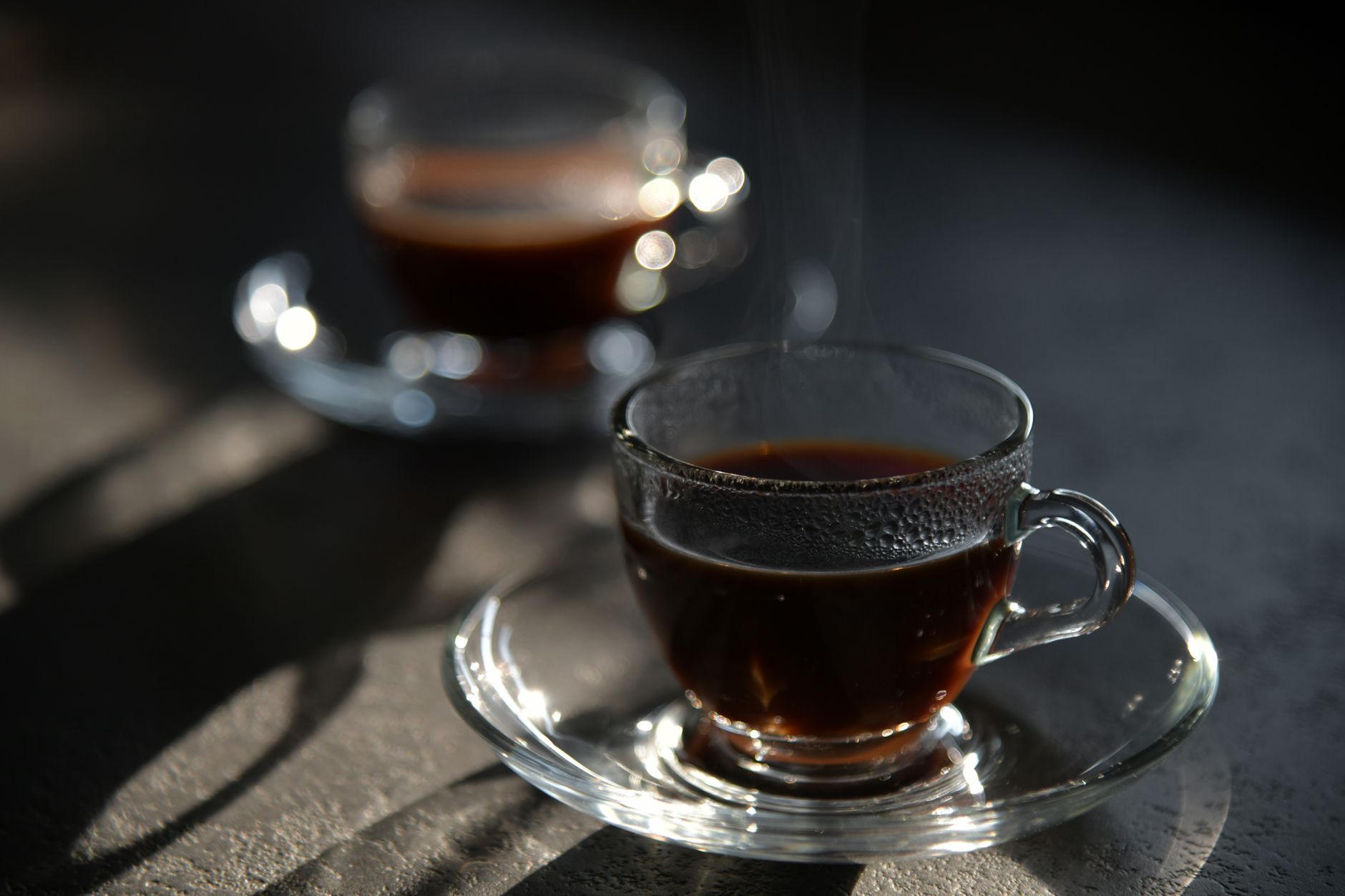 Primer plano de un café negro humeante en una elegante taza de vidrio con platillo, captado con luz suave.