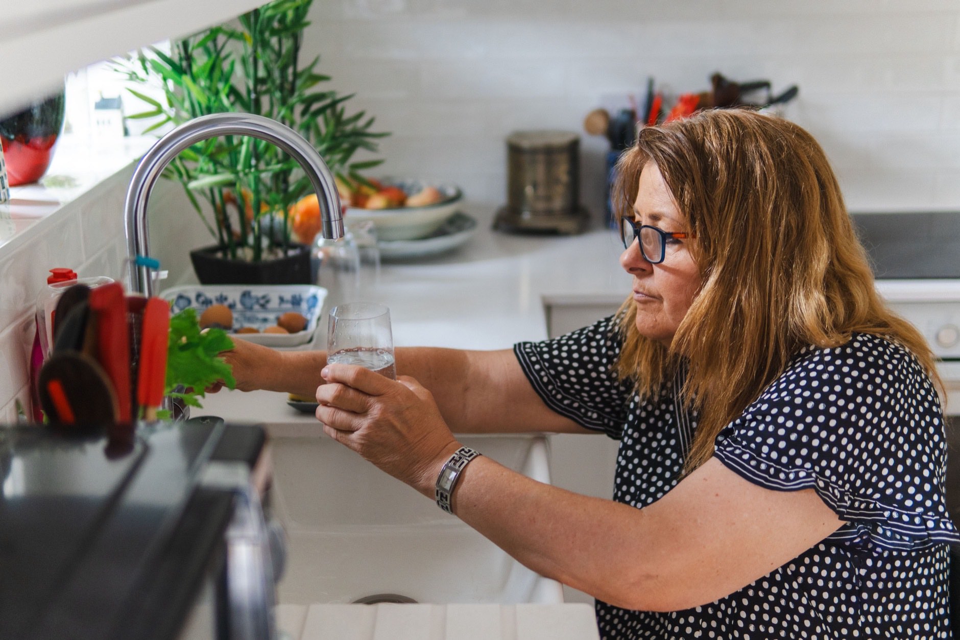 Mujer de mediana edad sirviéndose un vaso de agua en una cocina luminosa con verduras y fruta fresca