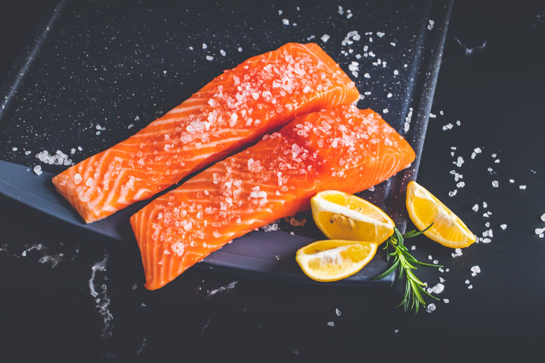 Primer plano de filetes de salmón crudo con sal, rodajas de limón y romero sobre una bandeja para hornear.