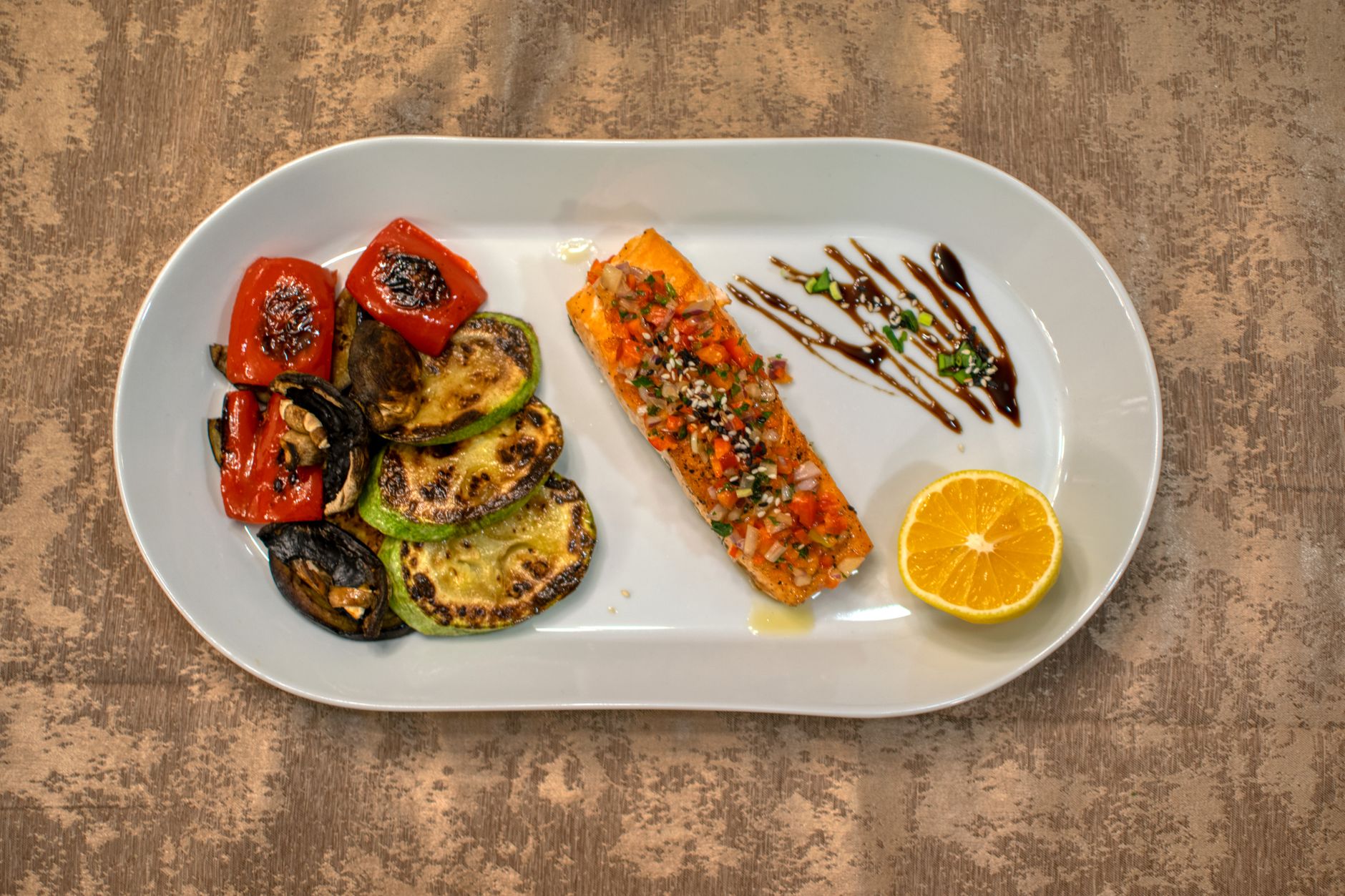 Salmón a la parrilla con verduras asadas y una rodaja de limón en un plato blanco.